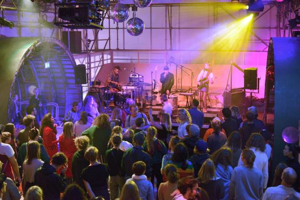 Eine Band spielt vor Publikum auf einer Bühne mit Discokugeln und farbiger Beleuchtung.