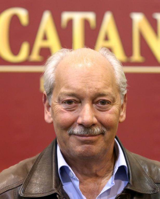 Klaus Teuber, der Erfinder des Brettspiels „Die Siedler von Catan“, porträtiert vor dem Catan-Logo.