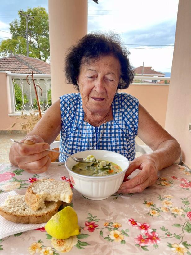 Eine ältere Frau isst Suppe mit Brot und Zitrone auf einem blumigen Tisch im Freien.