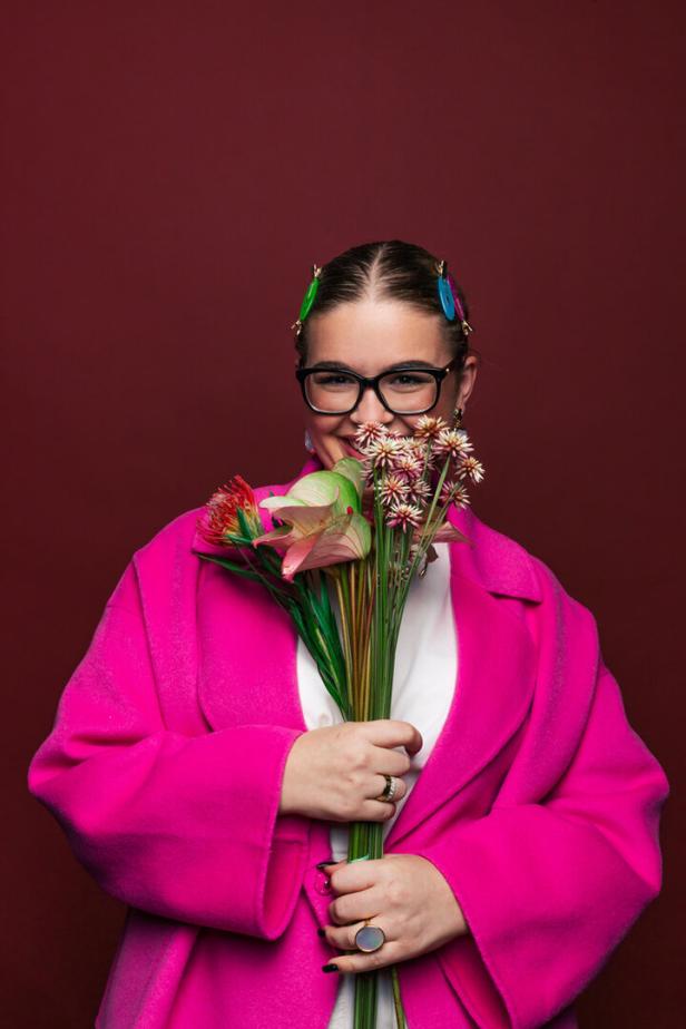 Eine Frau mit Brille und pinkfarbenem Mantel hält einen Blumenstrauß vor einem weinroten Hintergrund.