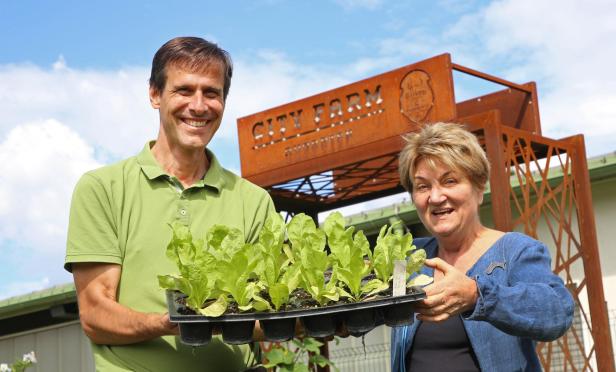 Zwei Personen halten Setzlinge vor einem Schild mit der Aufschrift „City Farm“.