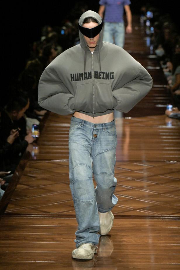 Ein Model mit futuristischer Brille und übergroßem Hoodie mit der Aufschrift „Human Being“ auf einem Laufsteg.