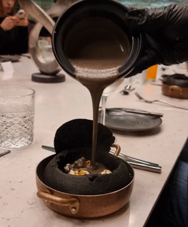 Eine dunkle Suppe wird in ein schwarzes Brot gegossen, das in einem Topf mit goldenen Griffen steht.