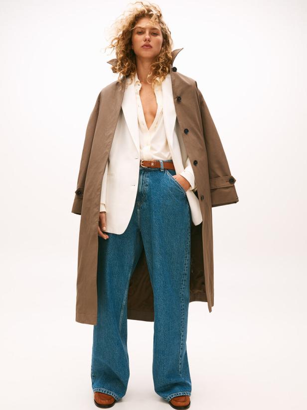 Eine Frau mit lockigem Haar trägt einen Trenchcoat, eine Bluse und weite Jeans.