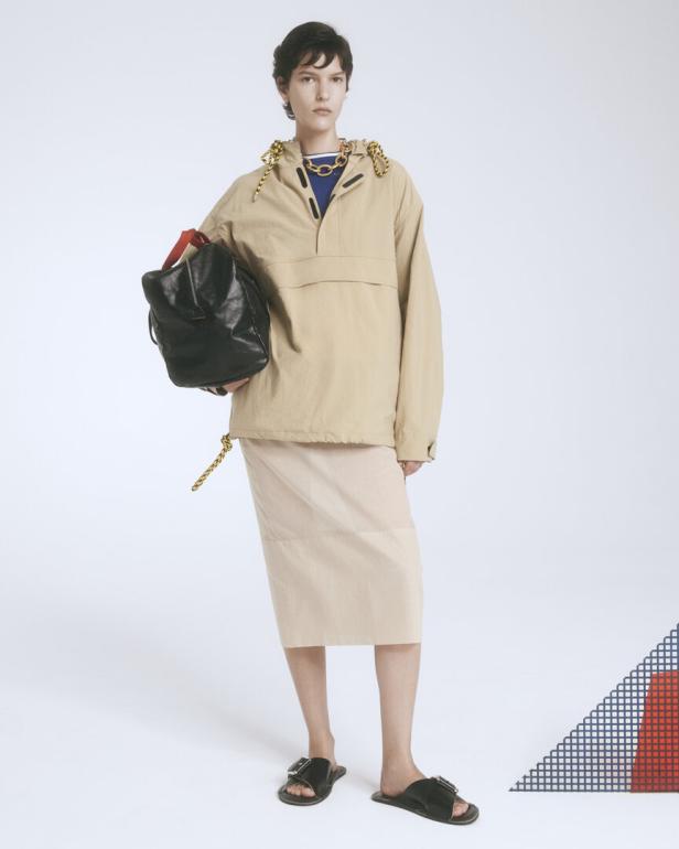 Eine Frau mit kurzer brauner Haaren trägt eine beige Jacke, einen Rock und schwarze Sandalen.