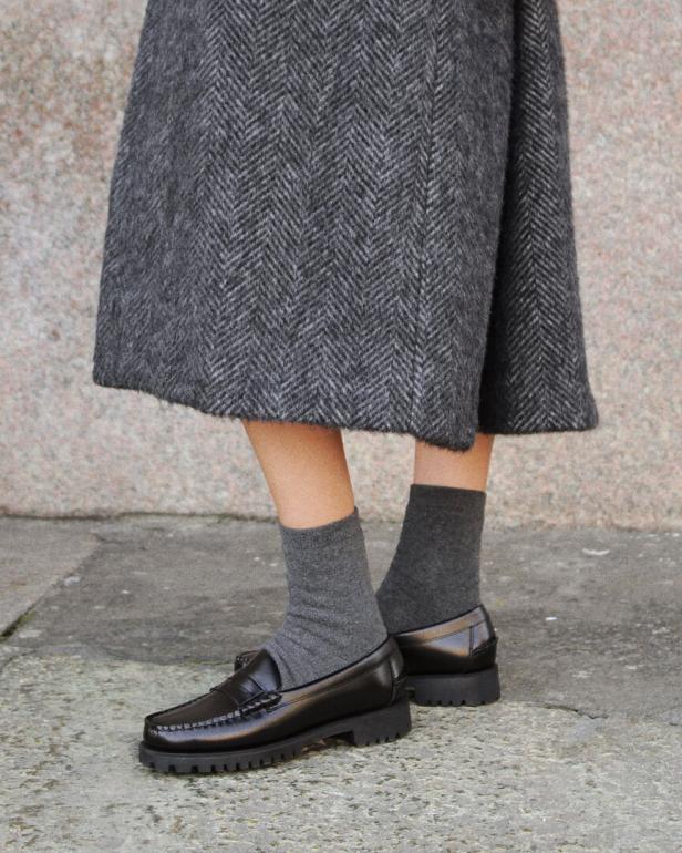 Nahaufnahme einer Person mit grauem Rock, grauen Socken und schwarzen Loafern.