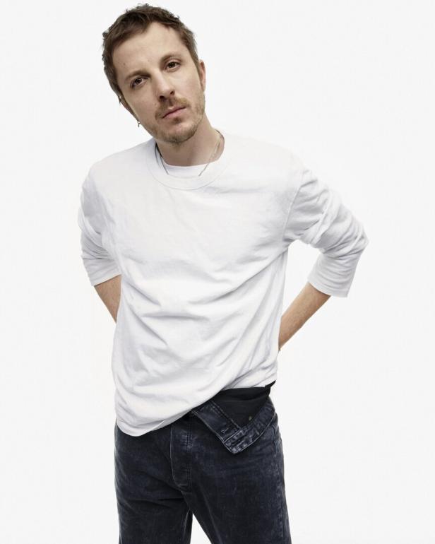 Ein Mann mit weißem Langarmshirt und dunkler Jeans posiert vor einem weißen Hintergrund.