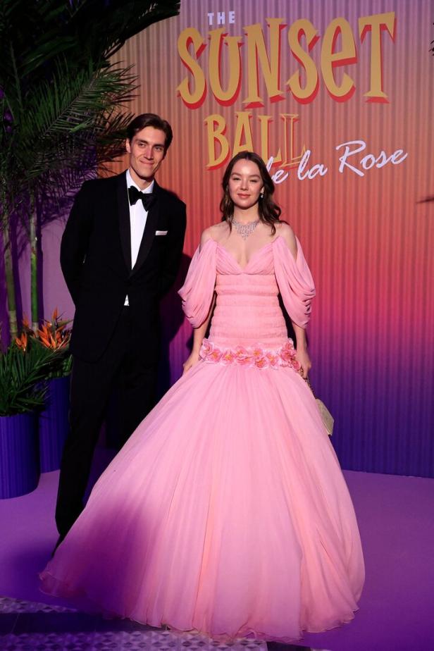 Ein Mann im Smoking und eine Frau in einem rosa Ballkleid posieren vor dem „The Sunset Ball de la Rose“-Banner.