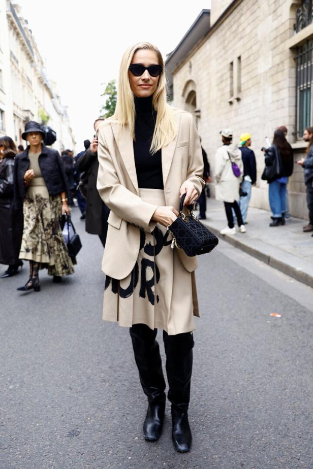 Eine Frau mit Sonnenbrille und beigem Blazer und Rock mit „Dior“-Aufdruck auf der Straße.