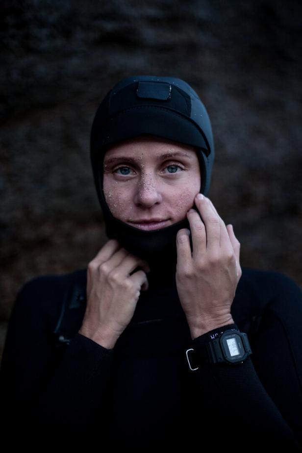Eine Frau mit Sand im Gesicht trägt einen schwarzen Neoprenanzug und eine Taucherhaube.