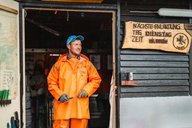 Ein Fischer steht in grell-orangen Klamotten in einer Holztür.