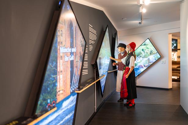 Zwei Personen in Tracht betrachten einen interaktiven Bildschirm in einem Museum.