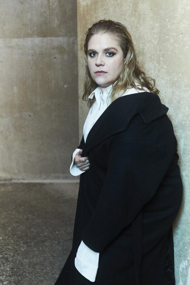 Eine Frau mit blonden, gewellten Haaren und dunklem Mantel posiert vor einer Wand.