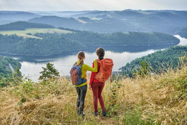 Zwei Frauen mit Rucksäcken stehen auf einem Hügel und blicken auf einen See und die umliegenden Berge.