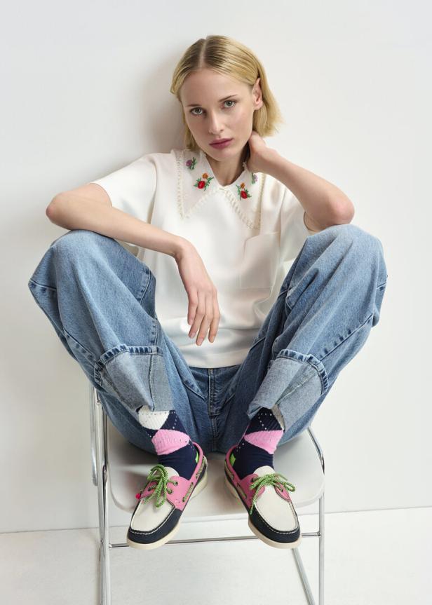 Eine Frau sitzt auf einem Stuhl und trägt eine weiße Bluse mit Blumenstickerei, Jeans und bunte Schuhe.