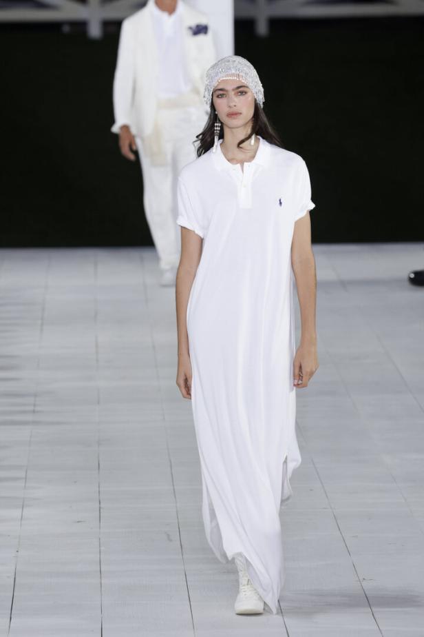 Eine Frau in einem weißen Polo-Kleid und einem glitzernden Kopfschmuck auf einem Laufsteg.