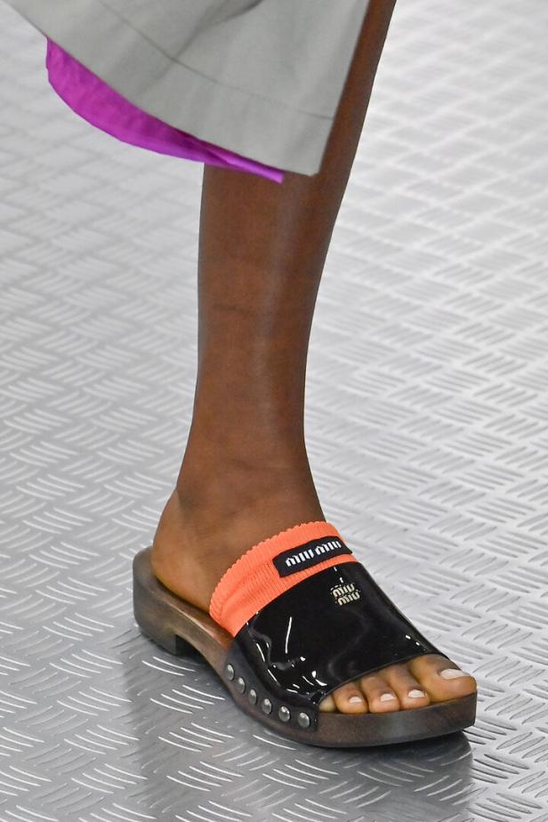 Nahaufnahme eines Fußes in einer Miu Miu Pantolette mit orangefarbenem Sockenabsatz.