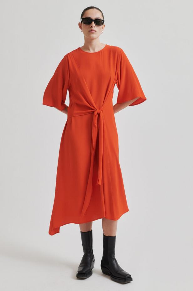 Eine Frau mit Sonnenbrille trägt ein orangefarbenes Kleid mit Wickeloptik und schwarze Stiefel.