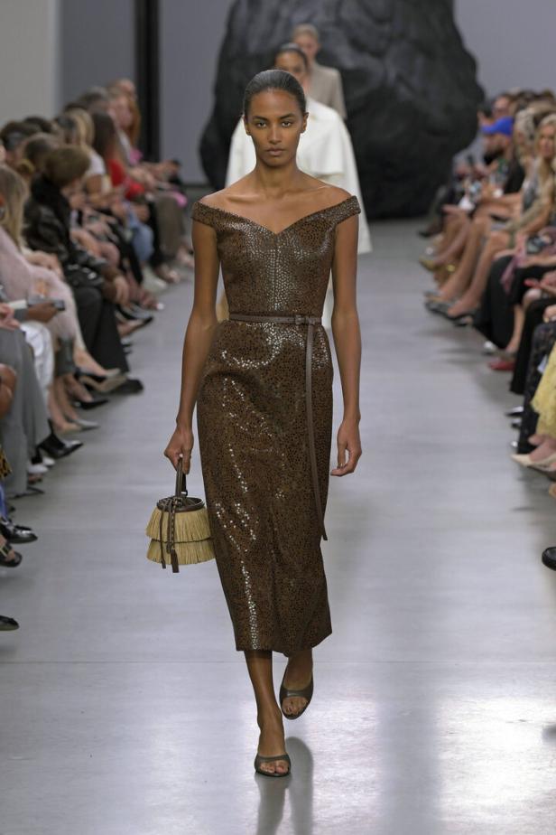 Eine Frau in einem braunen, schulterfreien Kleid mit Gürtel auf einem Laufsteg.