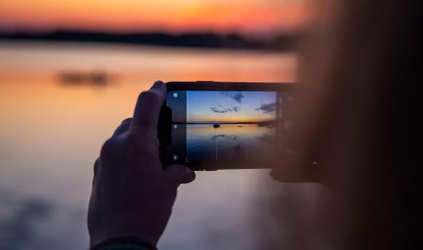 Eine Frau fotografiert mit ihrem Handy den sich spiegelnden Sonnenuntergang auf einem See.