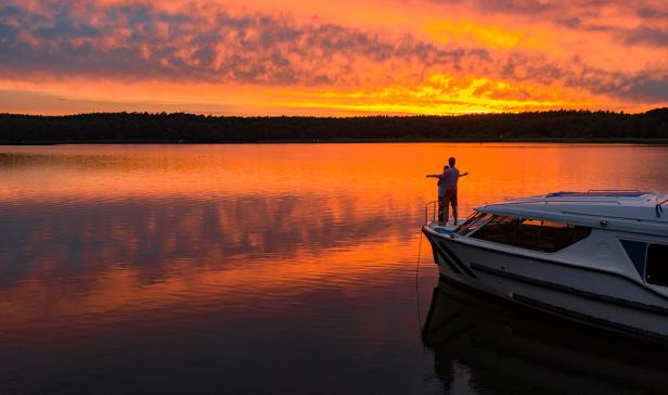 Ein Paar steht auf einem Boot in einem roten Sonnenuntergang, der sich auf einem See spiegelt.