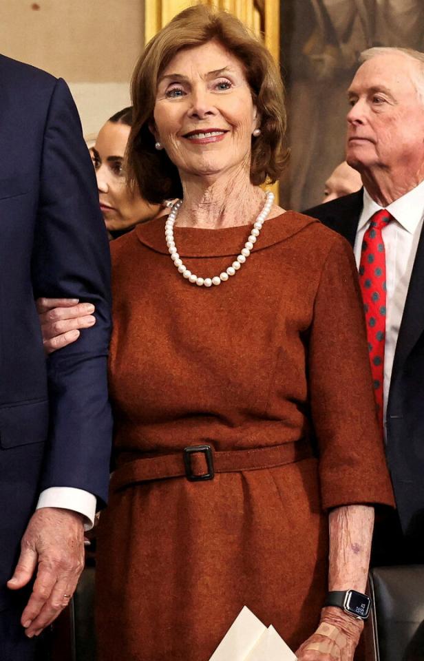 Eine ältere Frau mit braunem Kleid und Perlenkette steht zwischen zwei Männern.