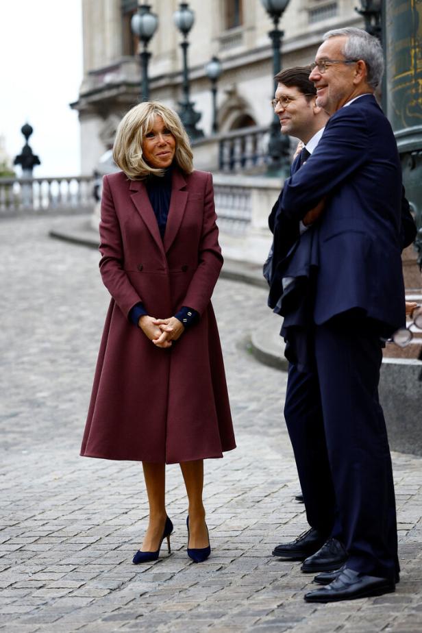 Brigitte Macron steht mit zwei Männern vor einem Gebäude.