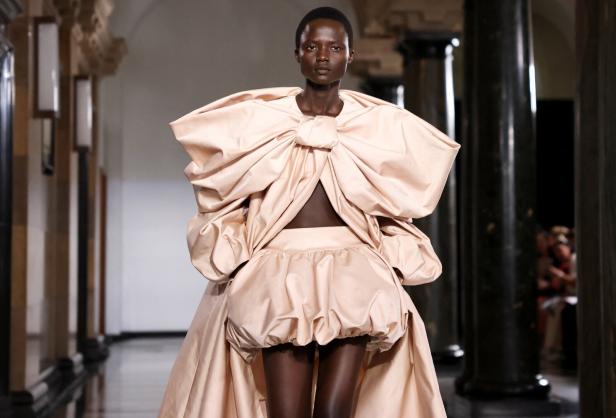 Eine Frau präsentiert ein extravagantes, hellrosa Kleid mit übergroßer Schleife auf dem Laufsteg.