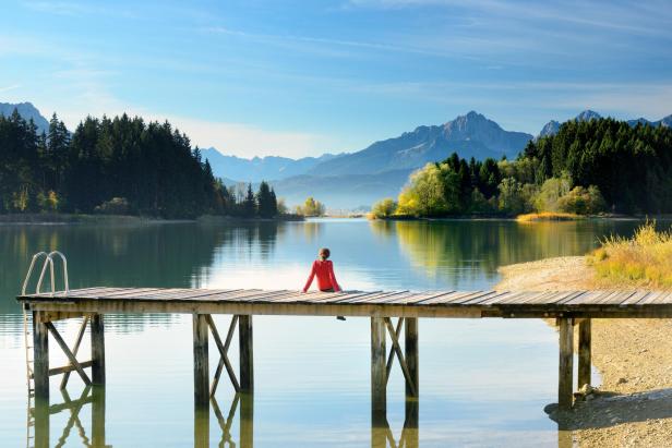 Ein Steg mit einer Person darauf, die an einem See sitzt und die Bergkulisse bewundert.