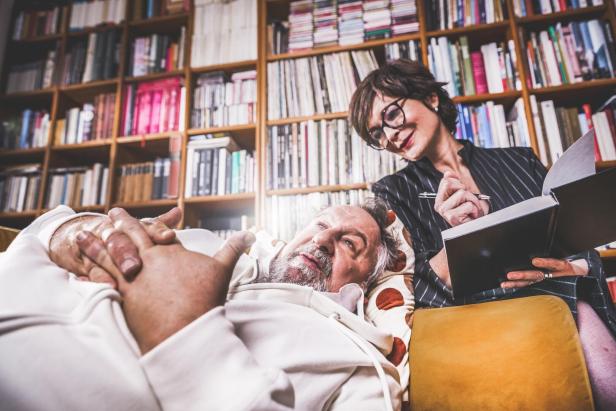Eine Frau liest einem liegenden Mann in einem Zimmer mit Bücherregalen vor.