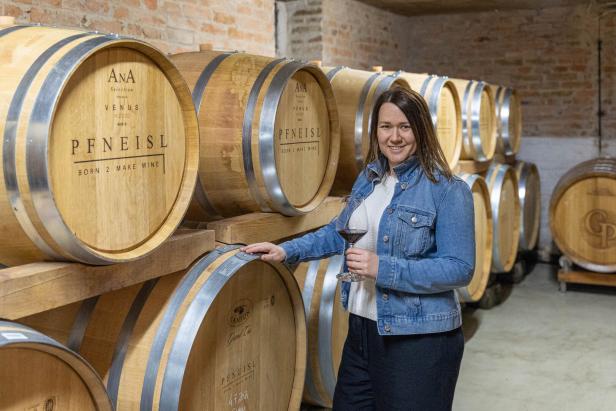 Eine Frau steht in einem Weinkeller mit Holzfässern und hält ein Glas Rotwein.