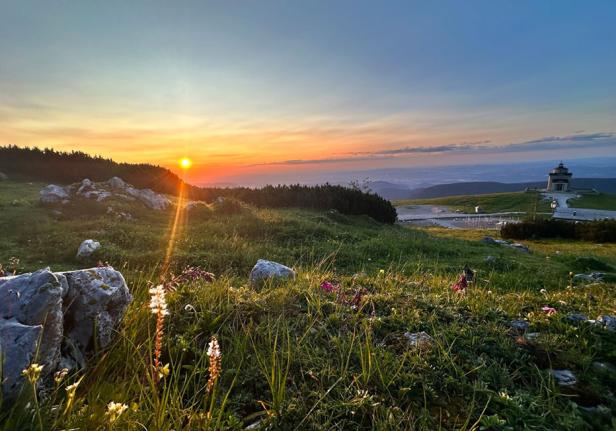 Sonnenaufgang über einer grünen Berglandschaft mit einem kleinen Gebäude im Hintergrund.
