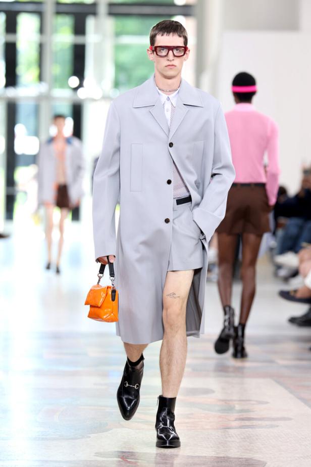 Ein Mann mit Brille und hellblauem Mantel läuft mit einer orangefarbenen Handtasche über einen Laufsteg.