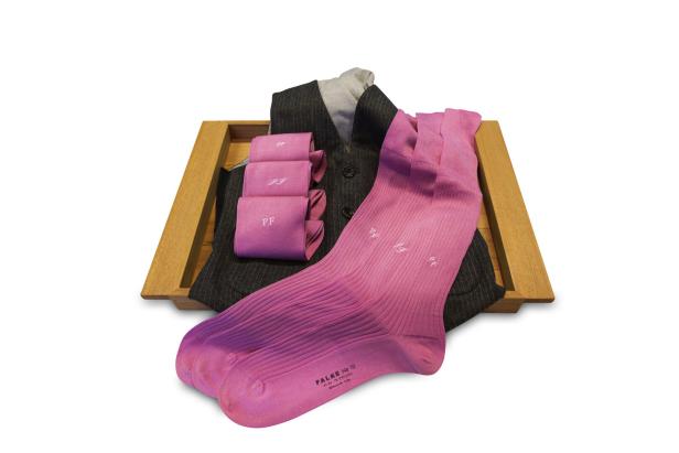 Ein Paar pinkfarbene FALKE-Socken, drei zusammengerollte, pinkfarbene Taschentücher und eine graue Weste auf einem Holztablett.