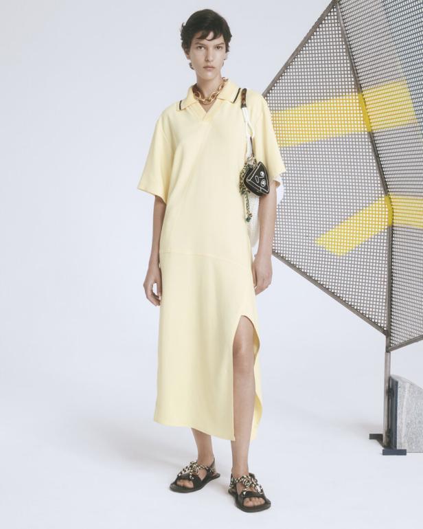Eine Frau in einem hellgelben Kleid mit Schlitz und Sandalen posiert vor einem Gitter.