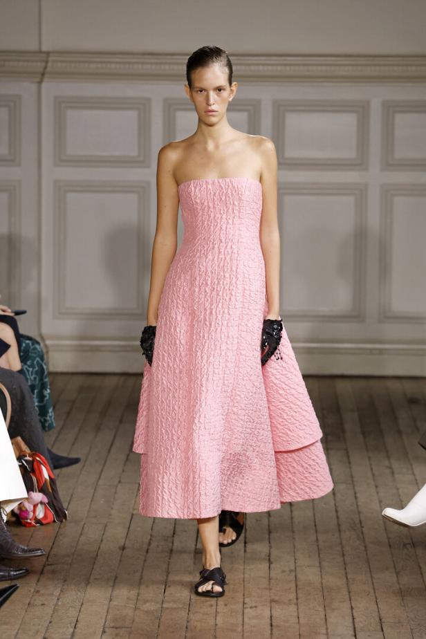 Ein Model präsentiert ein trägerloses, rosa Kleid mit strukturierter Oberfläche und schwarzen Sandalen auf einem Laufsteg.