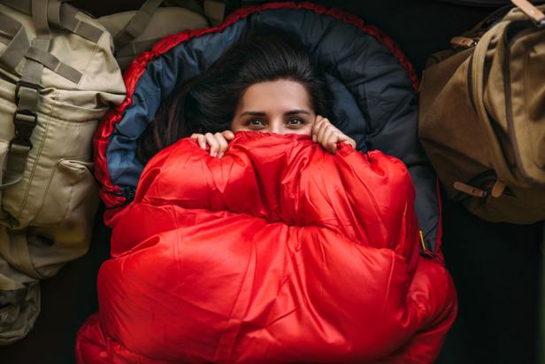 Eine Frau liegt in einem roten Schlafsack, umgeben von Rucksäcken.