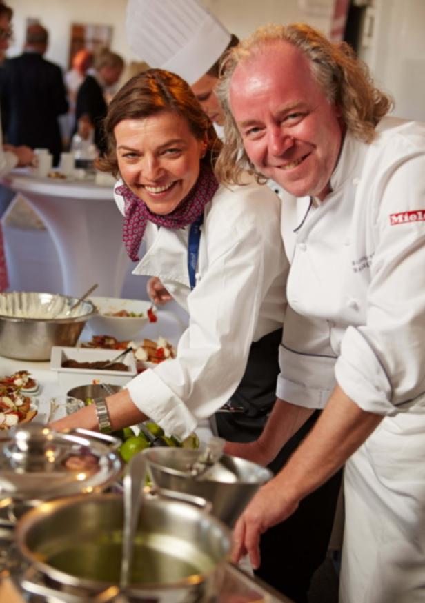 Zwei Köche bereiten Speisen in einer Küche zu und lächeln in die Kamera.