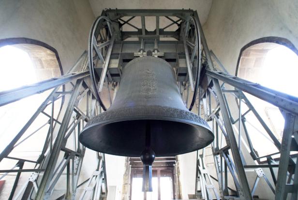 Eine große Glocke hängt in einem metallenen Glockenstuhl vor einem hellen Fenster.