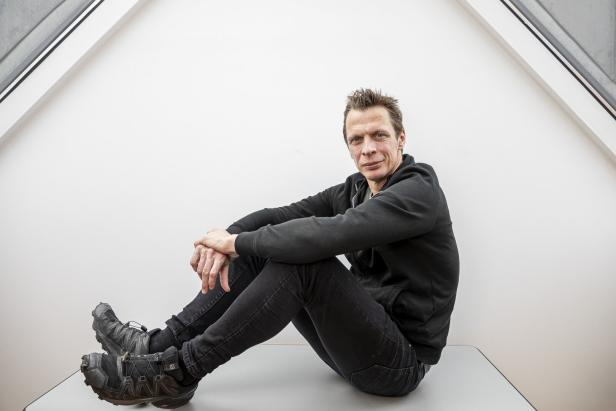 Ein Mann sitzt mit verschränkten Armen auf einem Tisch vor einer weißen Wand.