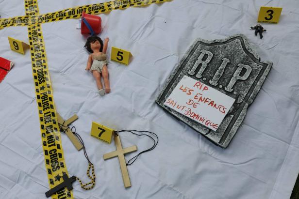FRANCE-EDUCATION-RELIGION-ASSAULT-INQUIRY-CHILDREN-DEMO Eine Puppe, Kreuze, ein Grabstein und Absperrband liegen auf einem weißen Tuch.