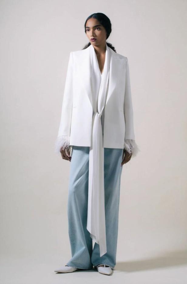 Eine Frau trägt einen weißen Blazer mit Federdetails und eine hellblaue Hose.