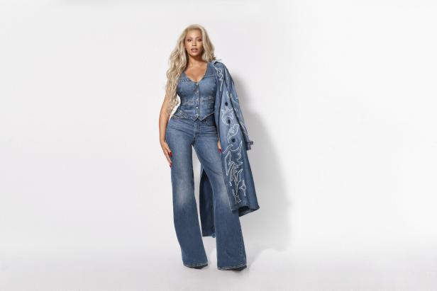 Eine Frau mit blonden, gewellten Haaren posiert in einem Jeans-Ensemble vor einem weißen Hintergrund.