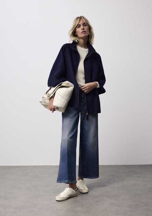 Eine Frau mit blonder Bob-Frisur trägt eine marineblaue Jacke, weite Jeans und weiße Turnschuhe.