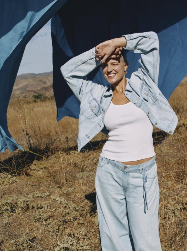 Eine Frau in Jeansjacke und Hose steht lächelnd in einer trockenen Landschaft.