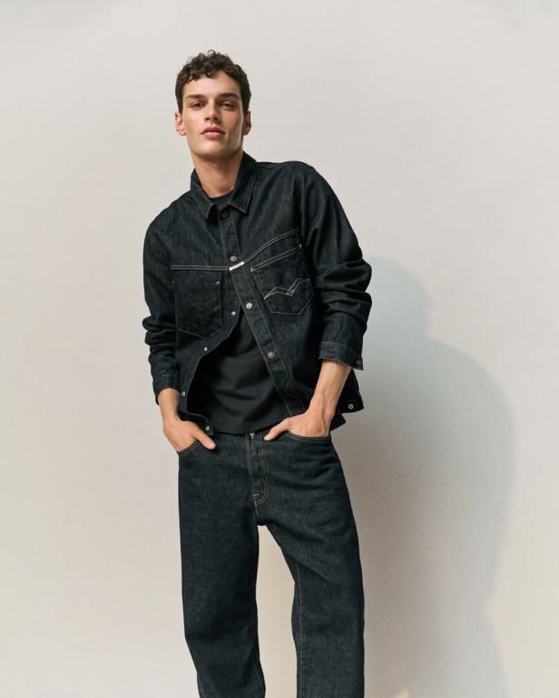 Ein Mann posiert in einem dunklen Jeanshemd und passender Hose vor einem hellen Hintergrund.