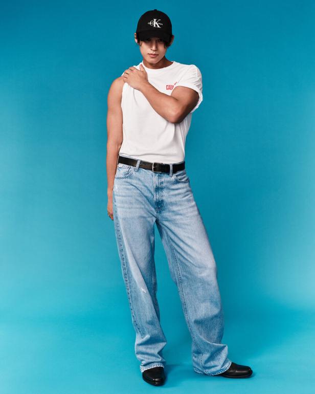 Ein Mann mit Kappe, T-Shirt und Jeans posiert vor blauem Hintergrund.