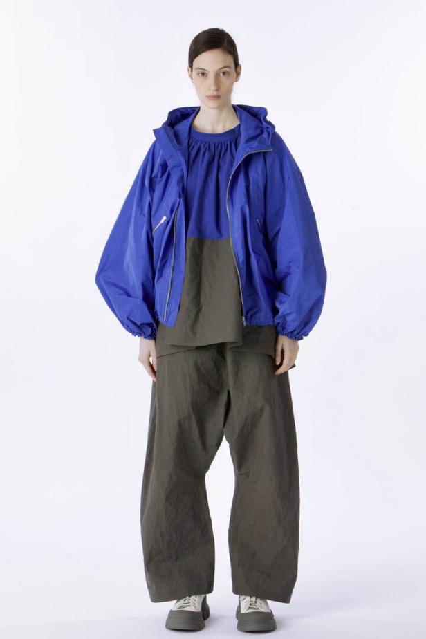 Eine Frau in einer blauen Jacke und weiten, olivgrünen Hosen vor einem weißen Hintergrund.