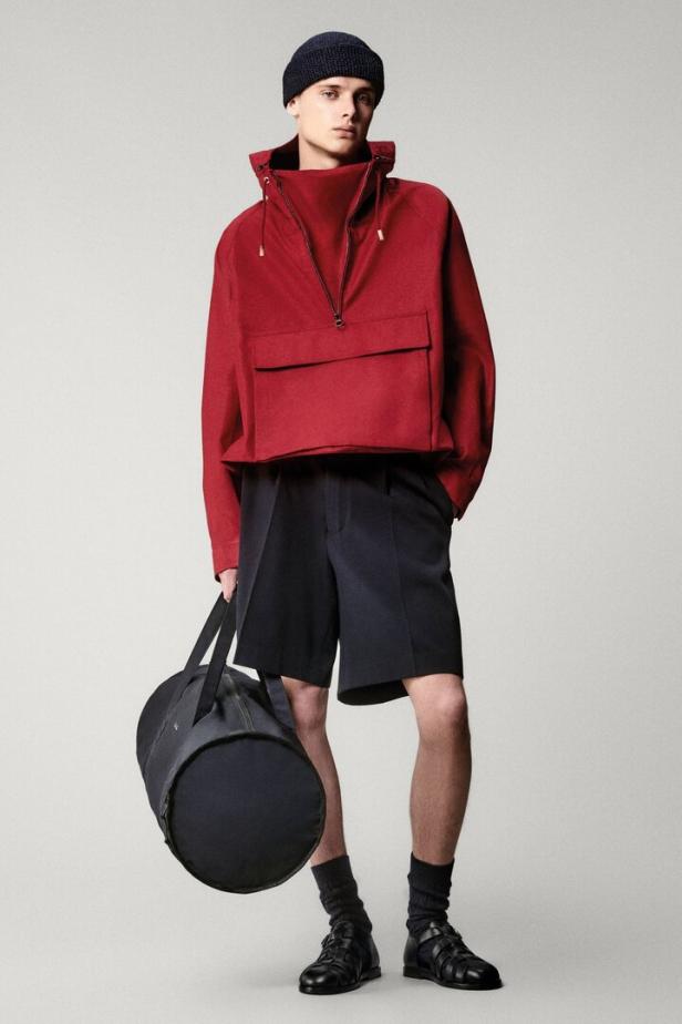 Ein Mann mit Mütze, rotem Anorak und Shorts hält eine Sporttasche.