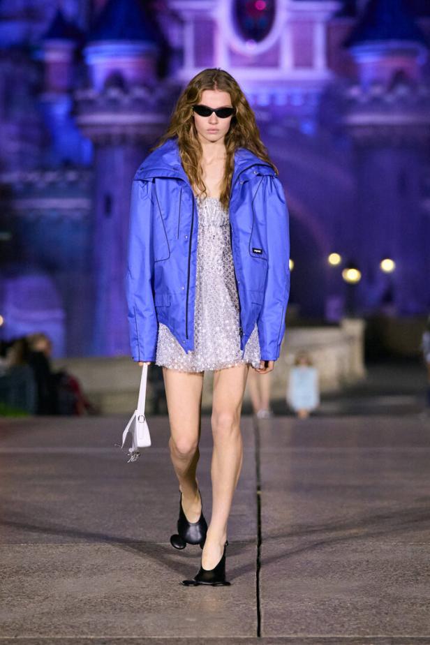 Eine Frau mit Sonnenbrille läuft in einem glitzernden Kleid und einer blauen Jacke vor einem Schloss.